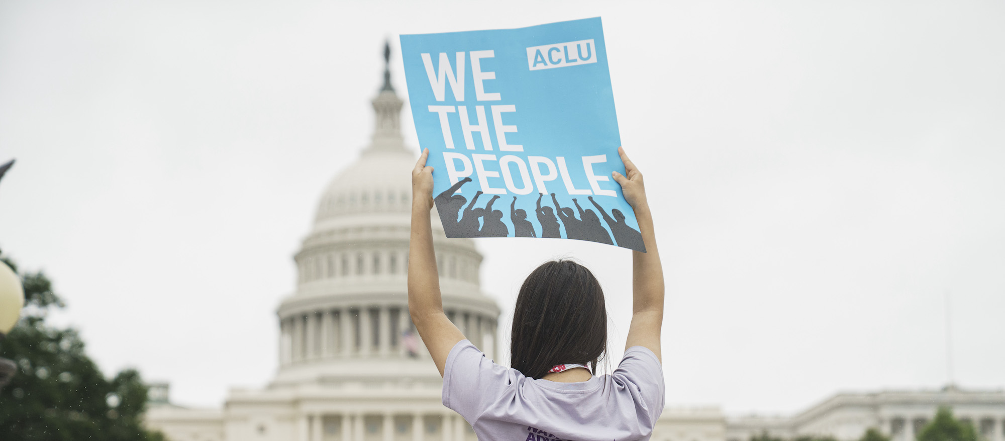 A woman holding a sign from the ACLU in front of the Capitol Building that says "we the people."