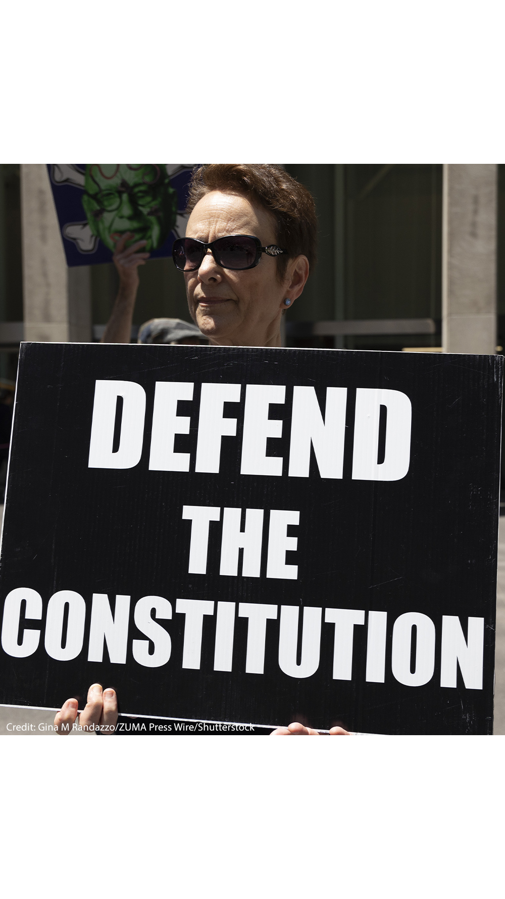 A demonstrator holds a sign (with white letters on a black background) that reads "DEFEND THE CONSTITUTION".