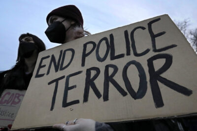 A demonstrator wearing a black hospital mask and black beanie hat holds up a cardboard sign that reads, "END POLICE TERROR".