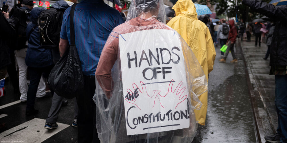 A person wearing a clear poncho displays a sign on their back that reads "HANDS OFF THE CONSTITUTION" while walking in the rain as they take part with thousands of other New Yorkers take part in the NO KINGS national day of action.