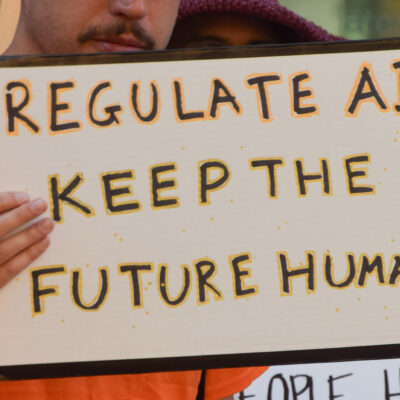 The camera focuses on a sign held by a demonstrator that reads, "REGULATE AI KEEP THE FUTURE HUMAN."