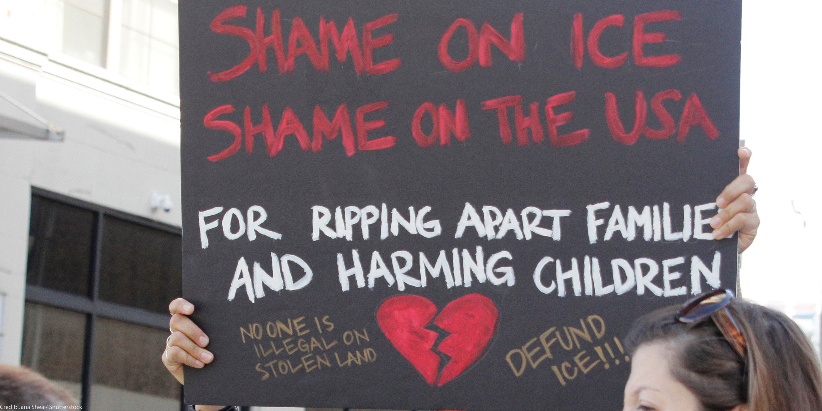 A demonstrator protests ICE and the Trump Administration policy of separating migrant families by holding up a sign (written in red ink on a black background with an illustrated broken red heart at the bottom) that reads, "SHAME ON ICE SHAME ON THE USA FOR RIPPIING APART FAMILIES AND HARMING CHILDREN No One is Illegal On Stolen Land, Defund ICE!!!"