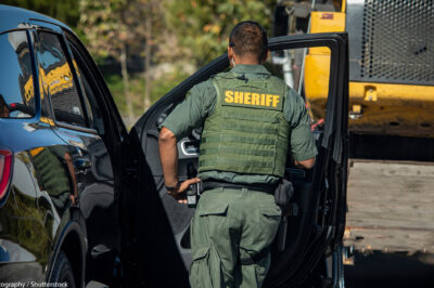 Shot of sheriff (wearing a bulletproof vest) from behind walking towards the open door of police car.