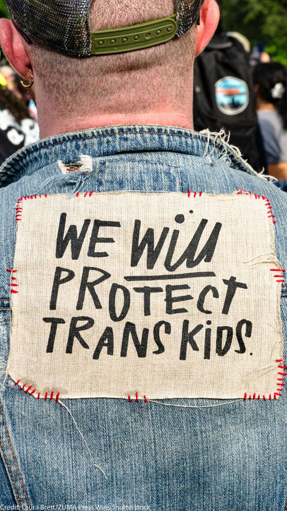 At a trans rally, person (wearing a denim vest and with their back to the camera) displays the sign stitched to the vest that reads "WE WILL PROTECT TRANS KIDS."